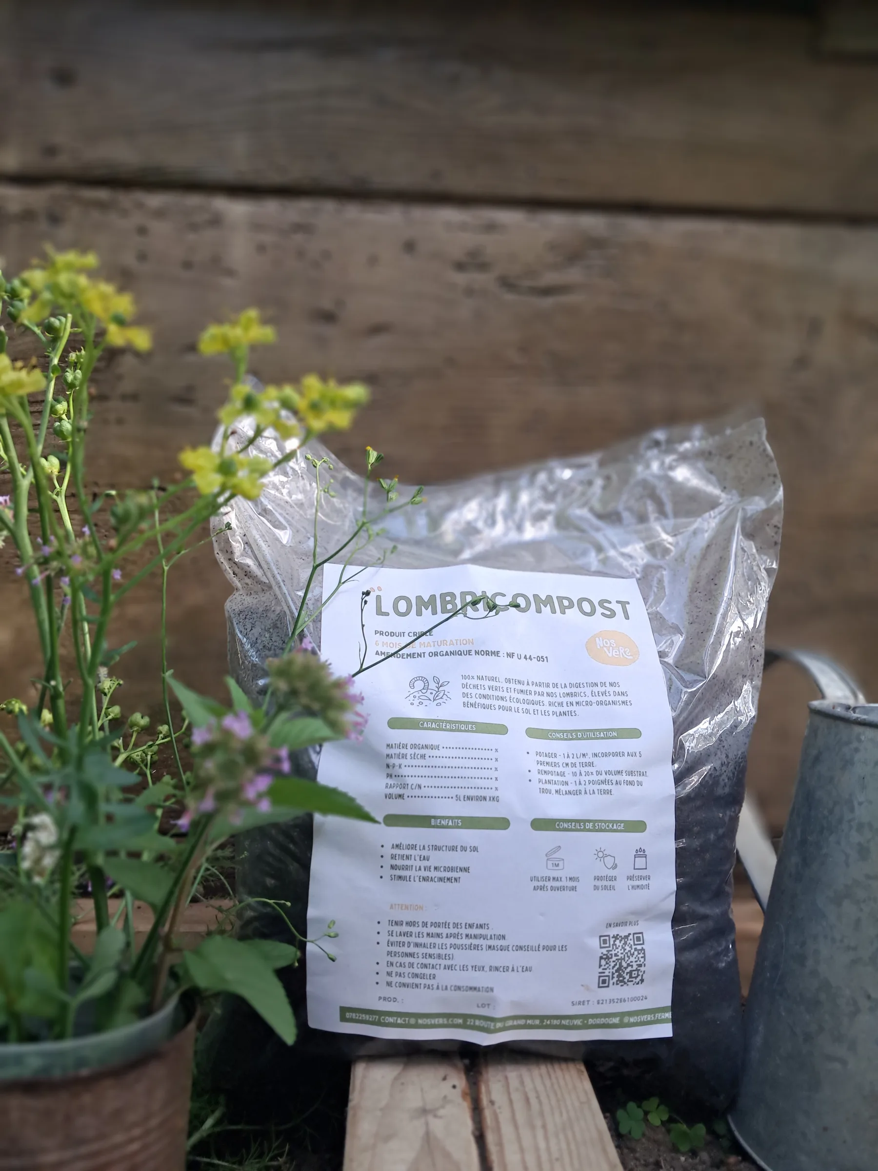 Un sac de lombricompost NosVers photographié à la ferme avec des fleurs et un arrosoir